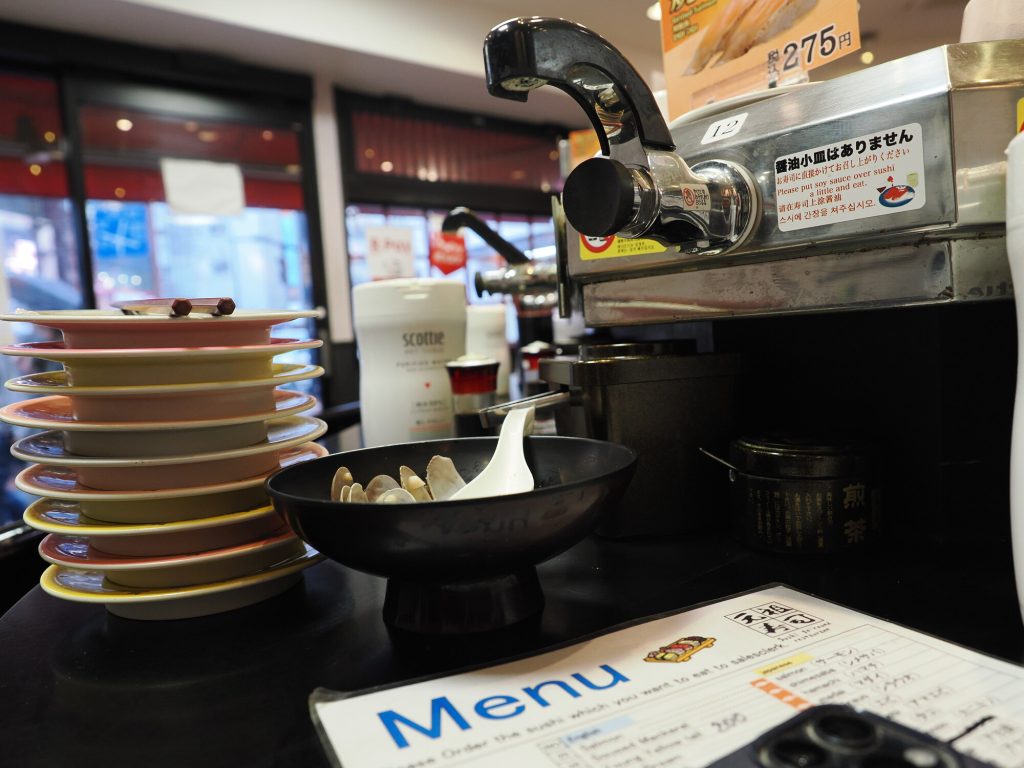 Stos pustych talerzyków po sushi w Ganso Zushi Ikebukuro - różowe żółte i szare talerze, kran sosu sojowego, miska z muszlami, menu na blacie