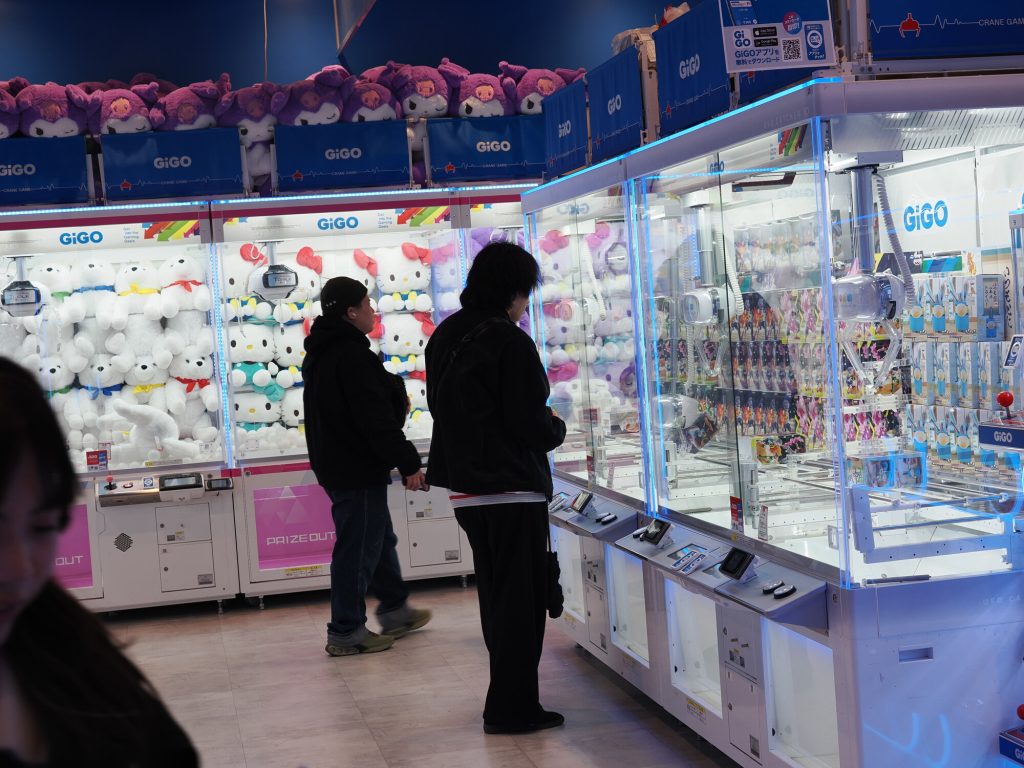 Dorośli gracze przy maszynach crane game w GiGO - dwóch mężczyzn analizuje maszynę z pluszakami Hello Kitty i figurkami, skupione spojrzenia