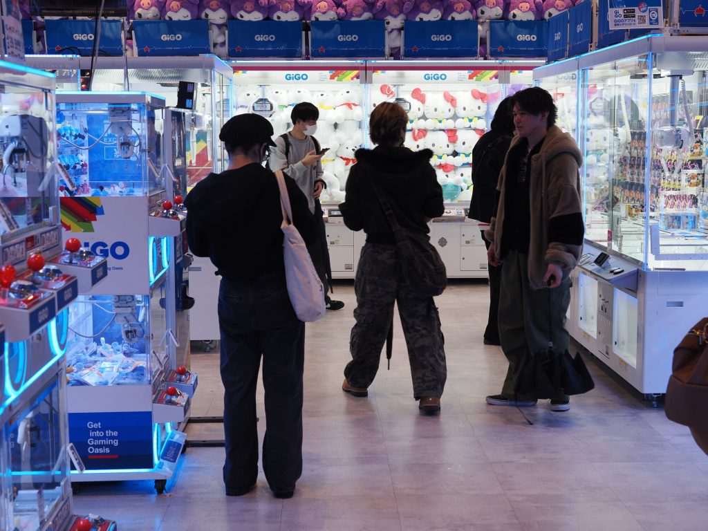 Korytarz w GiGO Ikebukuro - Get into the Gaming Oasis szyld, dorośli gracze przy maszynach, Hello Kitty pluszaki po sufitu, niebieski neon