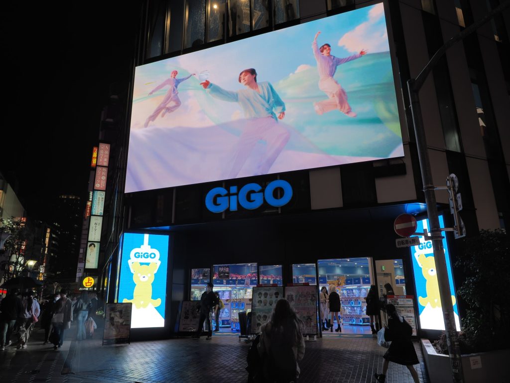 GiGO Ikebukuro nocna fasada - duży ekran LED z reklamą K-pop, niebieskie logo GiGO, żółty miś maskotka, wejście z maszynami widocznymi przez witrynę