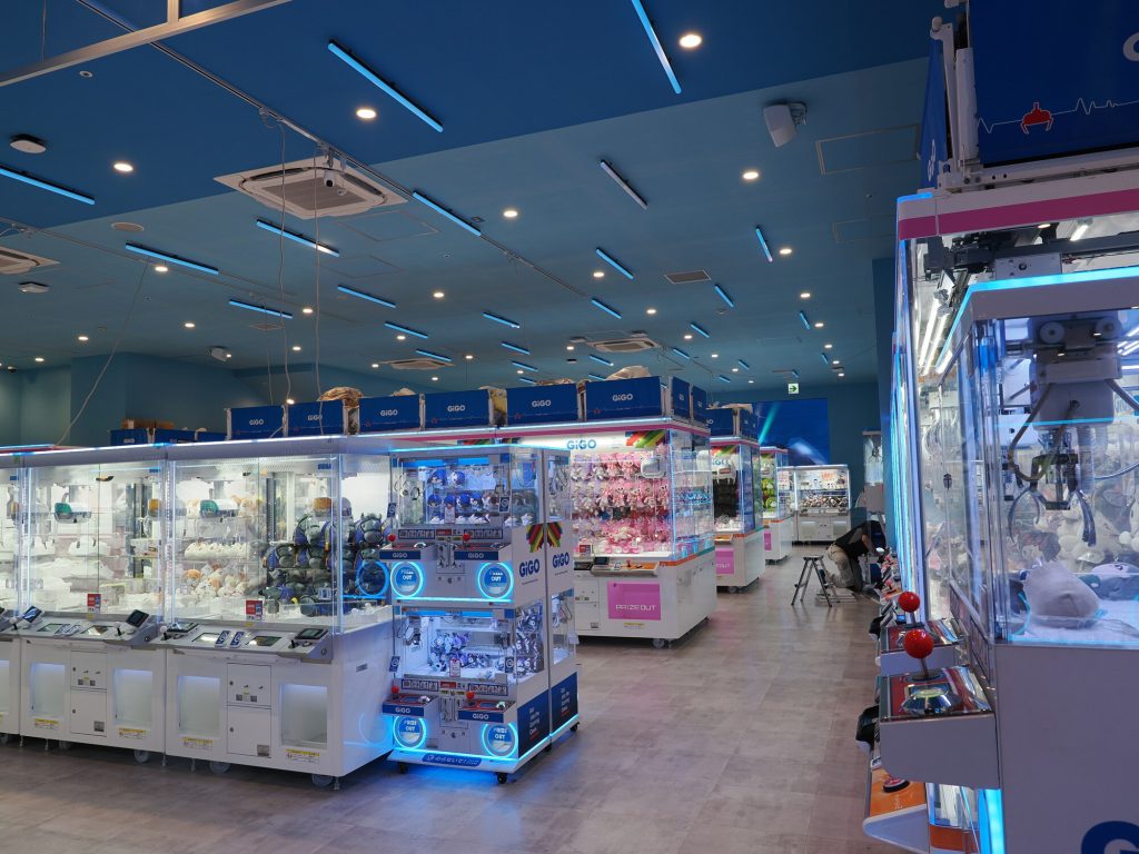 Wnętrze GiGO Ikebukuro - rzędy crane game maszyn w niebieskim oświetleniu, napisy PRIZE OUT, figurki i pluszaki w szklanych gablotach