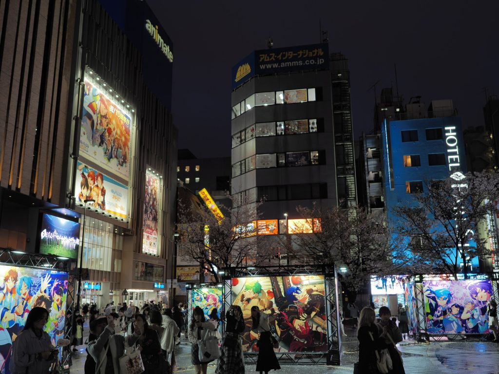 Plac przed Animate Ikebukuro nocą - plansze Ensemble Stars, tłum, logo Animate, drzewa sakury bez liści