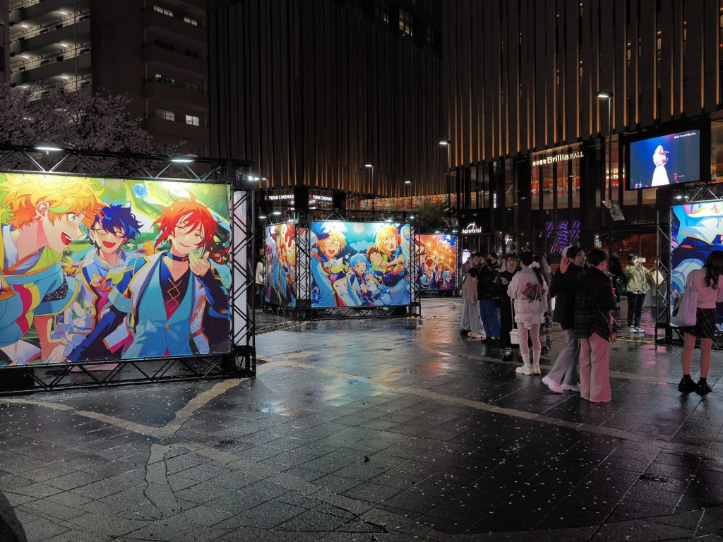 Plac przed Brillia Hall w Ikebukuro nocą - podświetlone plansze z anime Ensemble Stars na ruszowaniach, fangirls przy plakatach