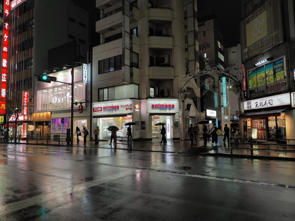 Animega Softmap w Ikebukuro nocą - biało-niebieski szyld アニメグッズ専門店 TAX FREE, mokry chodnik, przechodnie z parasolami