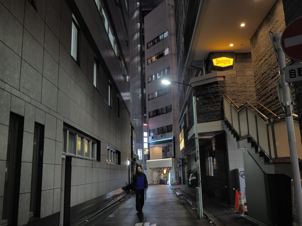 Boczna aleja w Ikebukuro nocą - wąska uliczka między wysokimi budynkami, sylwetka człowieka, Denny's szyld, mokra nawierzchnia