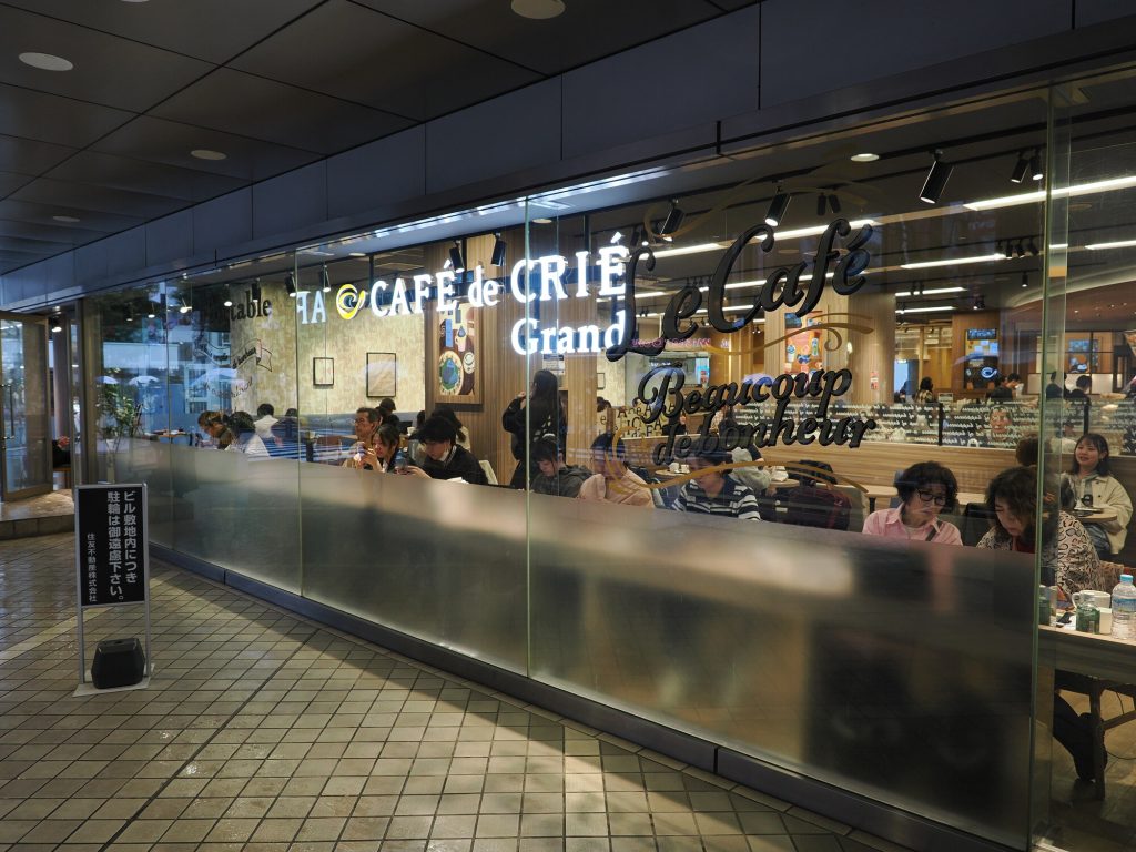 Café de Crié Grand w Ikebukuro nocą - przeszklona witryna pełna gości, napis Beaucoup de bonheur, Animate w tle