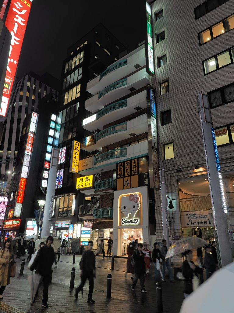 Nocne Ikebukuro - Sanrio sklep z neonem Hello Kitty na dole budynku, wyżej monjayaki i okonomiyaki, Sunshine City w tle