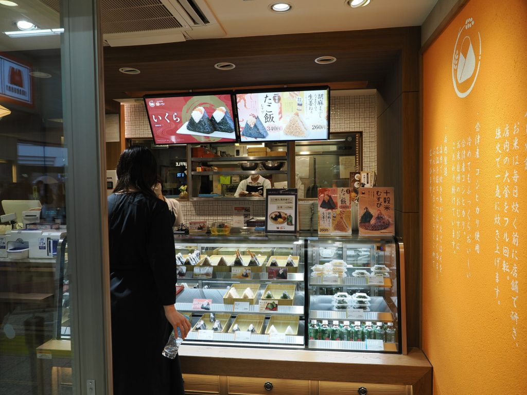 Sklep Musubi おむすび na stacji Ikebukuro - lada z onigiri w dwóch rzędach, szyldy いくら 340円 i たこ飯 340円, klientka wybierająca, pomarańczowa ściana z filozofią sklepu
