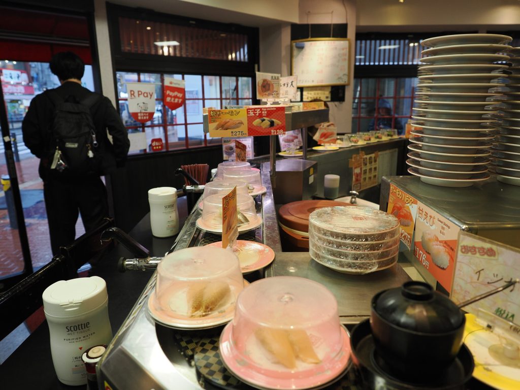 Taśma kaiten sushi przy wyjściu Ganso Zushi - R Pay i PayPay szyldy, deszcz za szybą