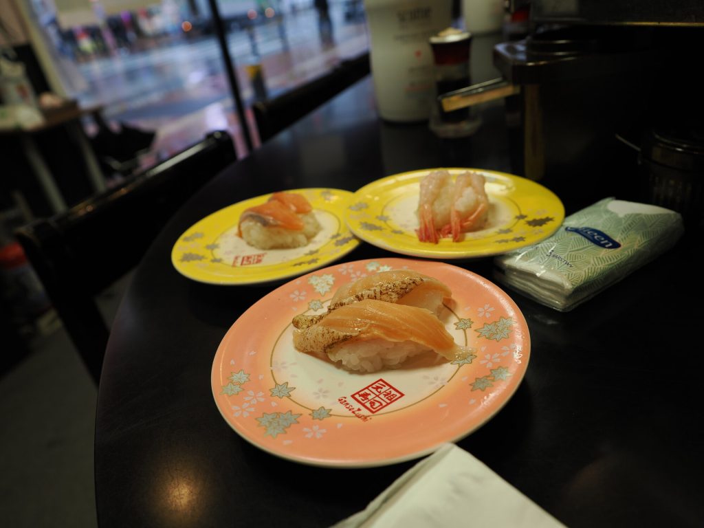 Trzy talerze sushi na stole Ganso Zushi - łosoś pstrąg krewetka słodka, okno z deszczem