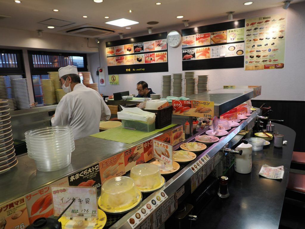 Wnętrze Ganso Zushi Ikebukuro - szef kuchni w białym czepku, starszy pan gość przy barze, taśma z talerzami