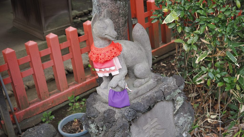 Kamienny posążek kitsune w czerwonym sweterku i fartuszku przy świątyni Hanazono Inari Jinja