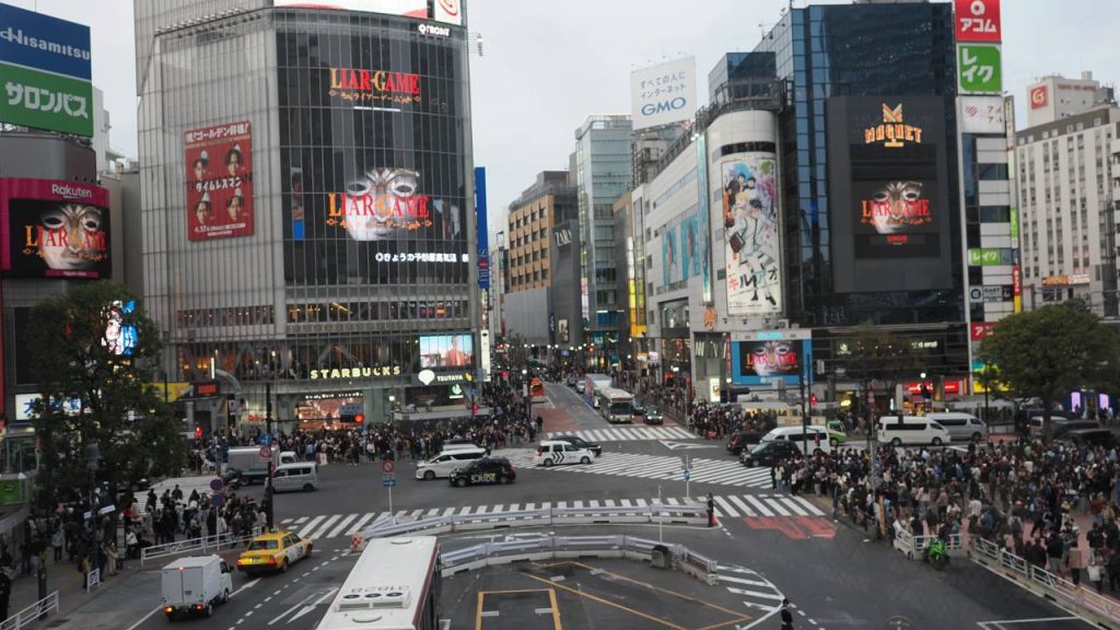 Shibuya Scramble Crossing z góry - światła czerwone, tłum czeka po wszystkich stronach, Starbucks i reklama Liar Game w tle