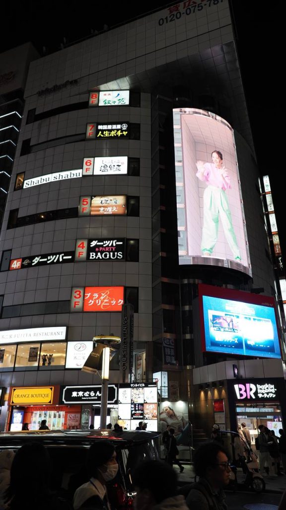 Zakrzywiony ekran 3D przy Shibuya z modelką w rozowym ubraniu, w tle budynek z shabu shabu dart bar BAGUS i klinika urody