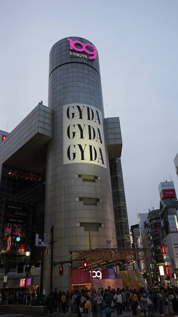 Budynek Shibuya 109 o zmierzchu - cylindryczna wieża z różowym logo 109, wielki napis GYDA GYDA GYDA, przed wejściem tłum