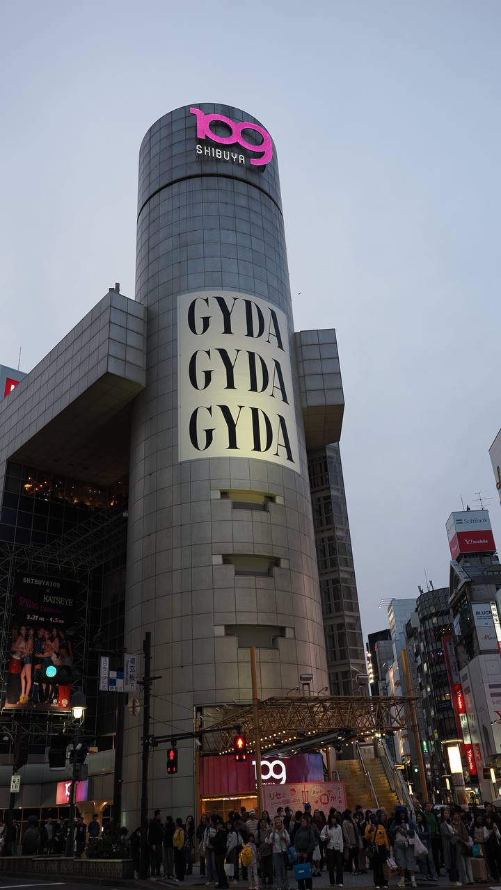 Budynek Shibuya 109 o zmierzchu - cylindryczna wieża z różowym logo 109, wielki napis GYDA GYDA GYDA, przed wejściem tłum