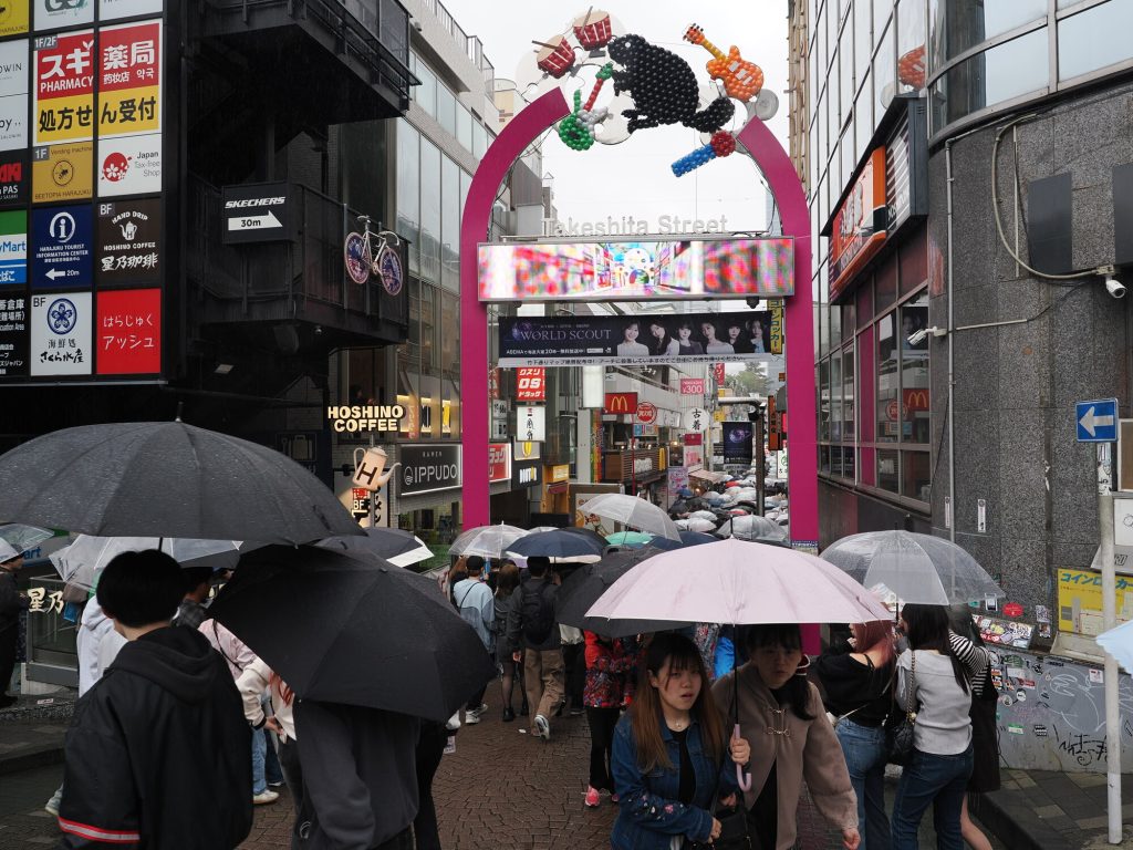 Wejście na Takeshita-dori - różowa brama z balonowym zwierzakiem, tłum parasoli, deszcz, baner World Scout