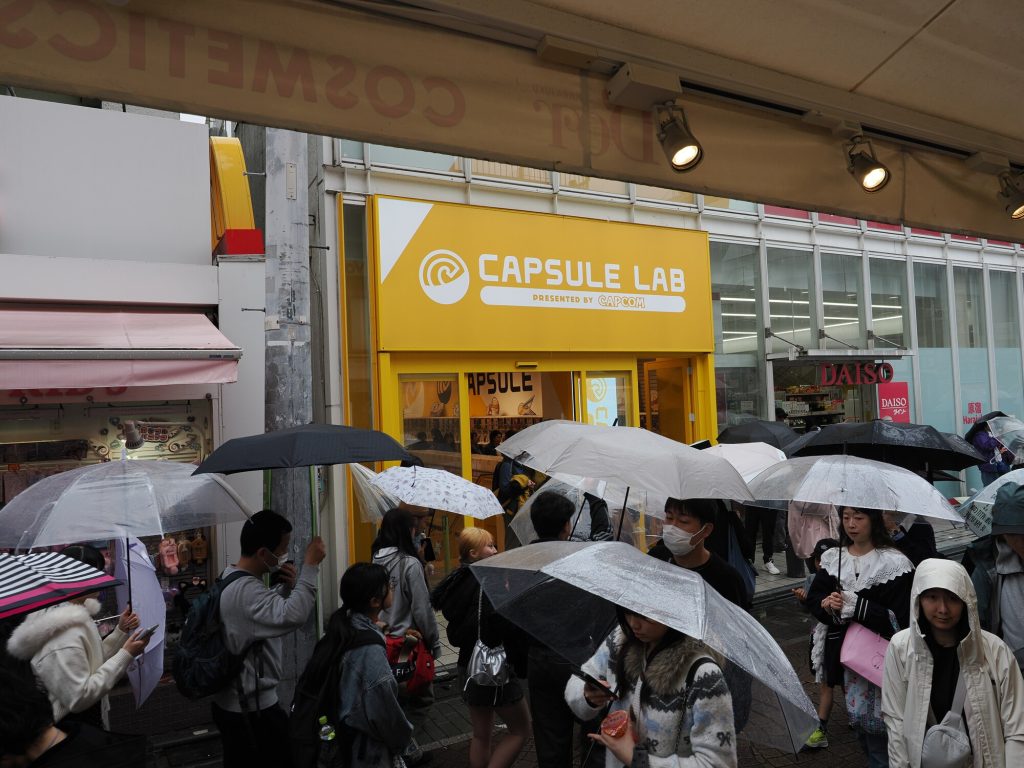 Wejście do Capsule Lab by Capcom na Takeshita-dori - żółty szyld, kolejka z parasolami w deszczu, Daiso w tle