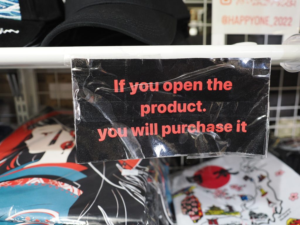 Karteczka na półce sklepu na Takeshita-dori: If you open the product. you will purchase it. W tle koszulki anime i czapki.