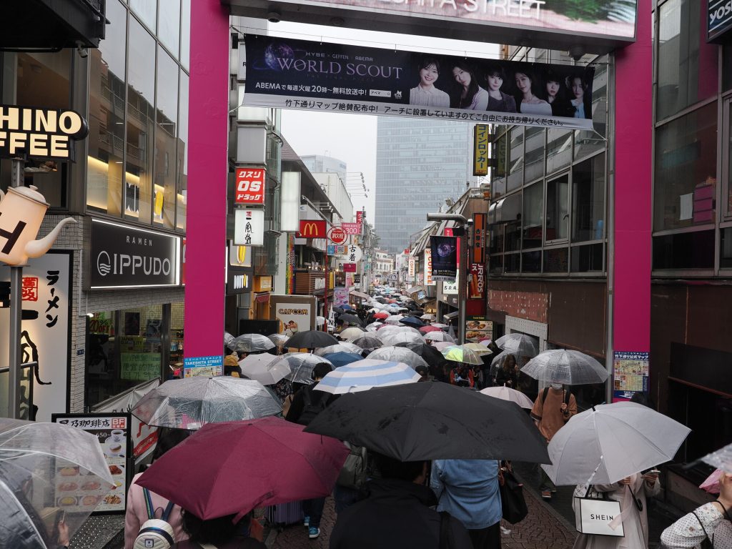 Widok wzdłuż Takeshita-dori przez różową bramę - morze parasoli, szyldy Ippudo Capcom McDonald's, wieżowiec w tle