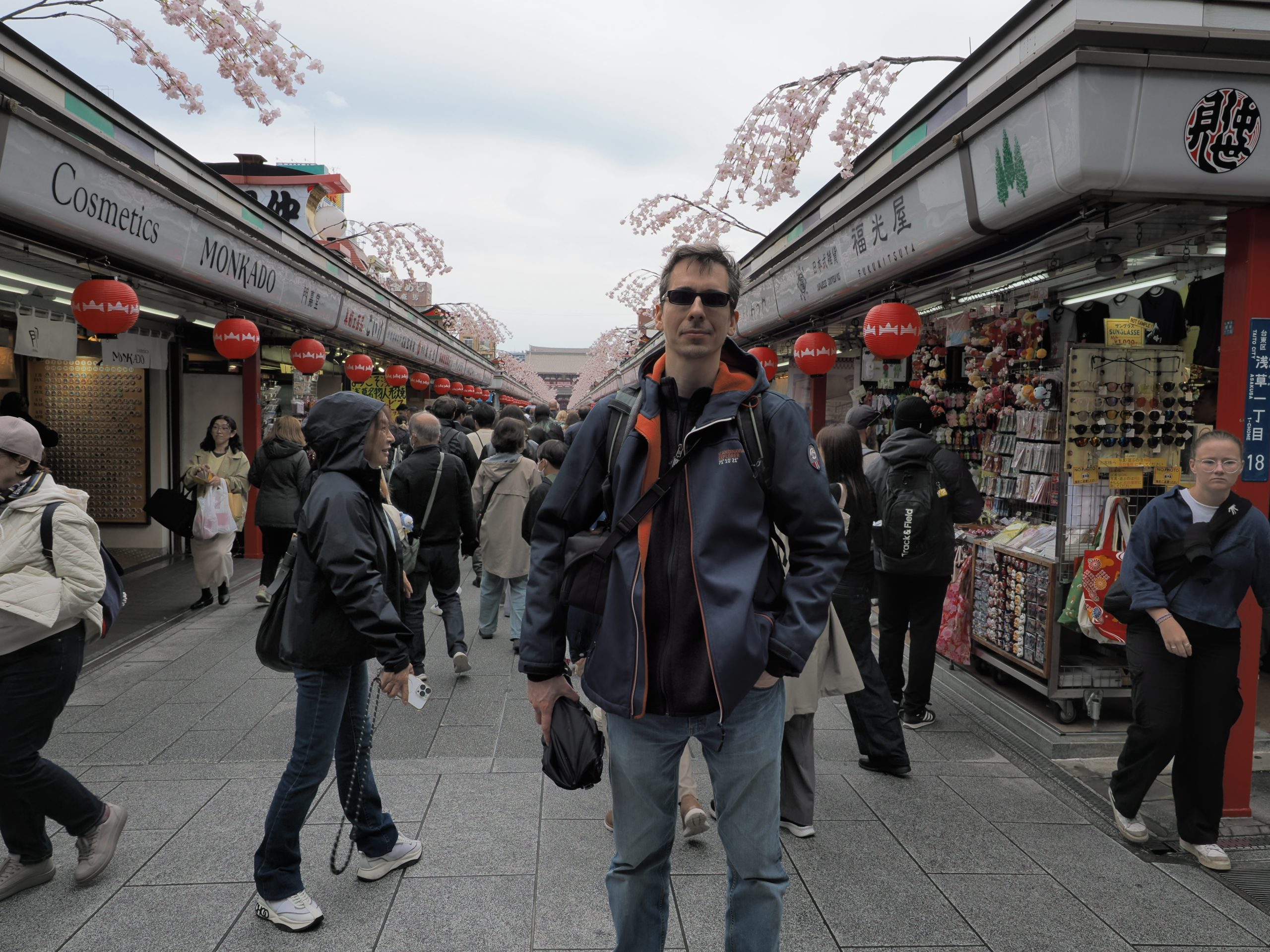 Kamil w okularach przeciwsłonecznych i niebiesko-pomarańczowej kurtce pozuje na Nakamise-dori, sakura nad głową