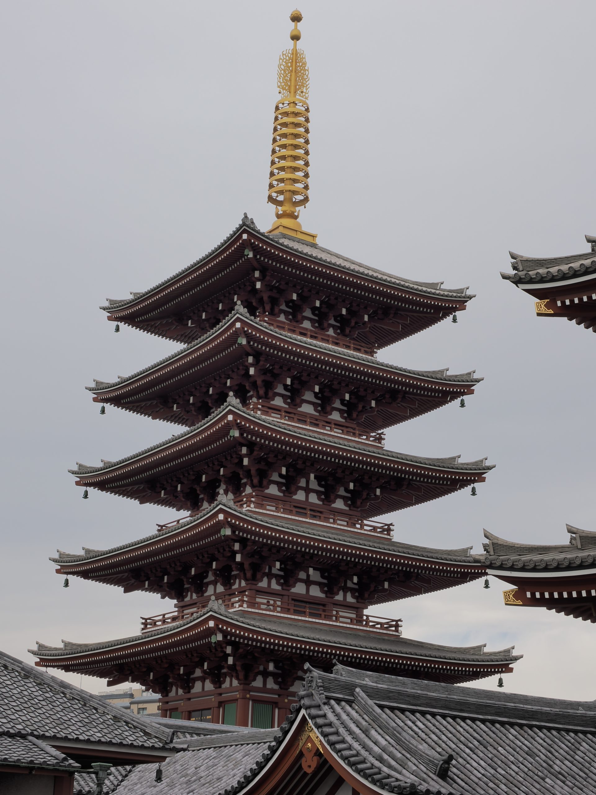 Zbliżenie pięciokondygnacyjnej pagody świątyni Senso-ji na tle szarego nieba, złoty finial na szczycie, ciemnoczerwona drewniana konstrukcja