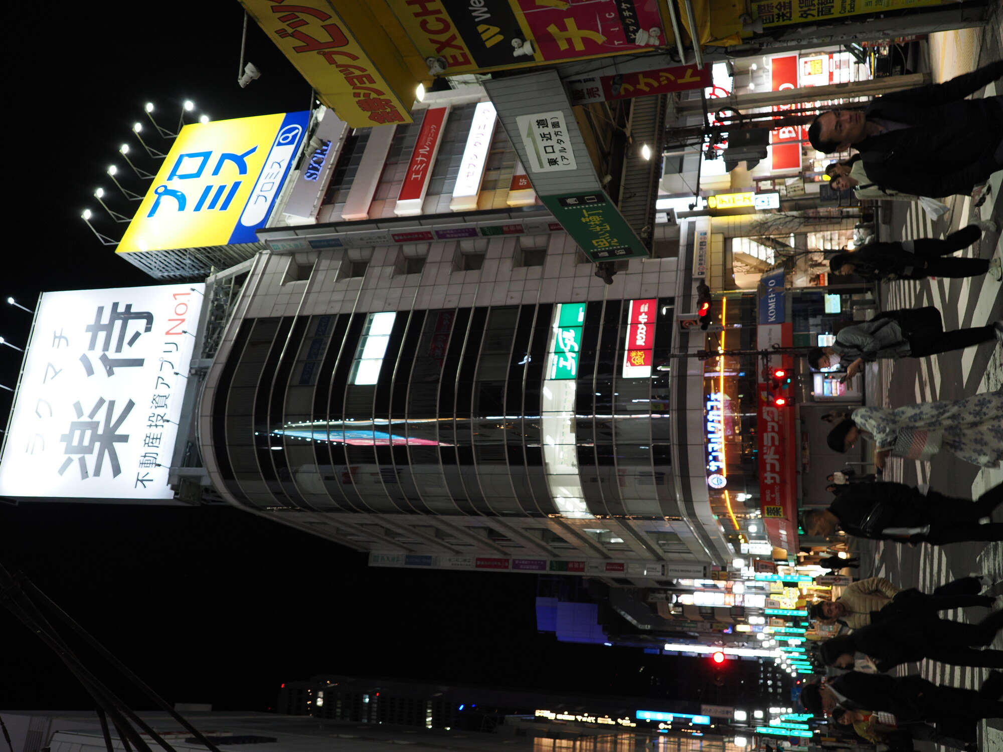 Nocne skrzyżowanie przy stacji Shinjuku — szyldy Rakumachi i Promise, tłum przechodniów