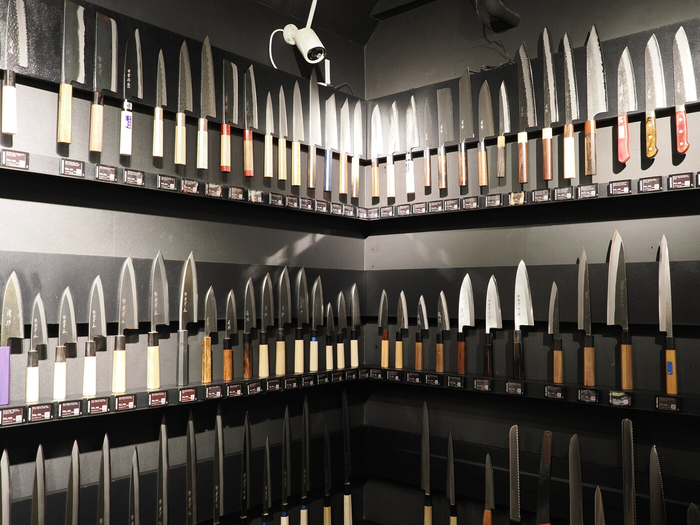 Ściana noży japońskich w sklepie — trzy poziomy półek z gyuto, yanagiba, nakiri i deba na czarnym tle