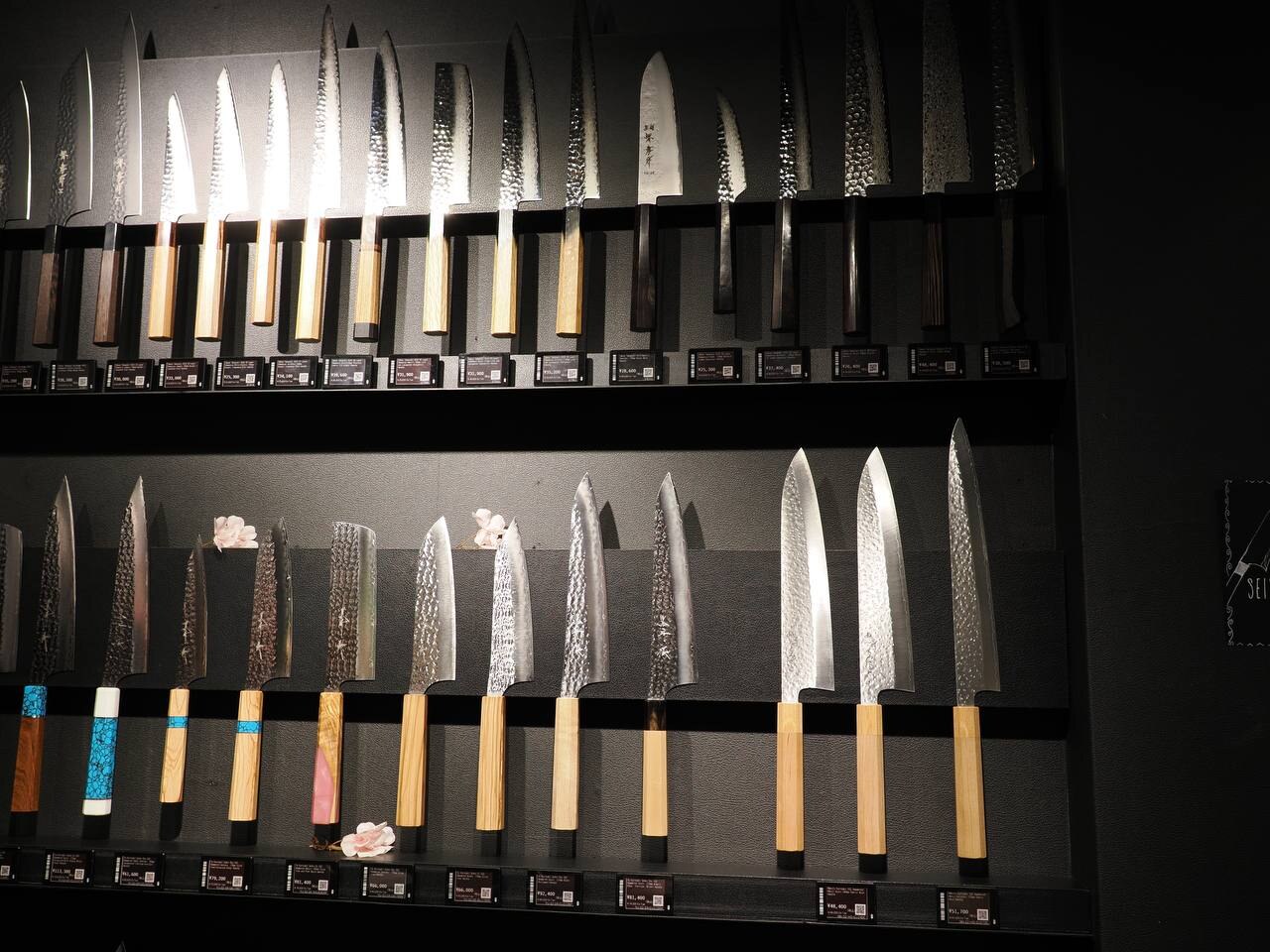 Wnętrze sklepu z nożami — ściany z setkami noży podświetlonych na czarnym tle, każdy z metką