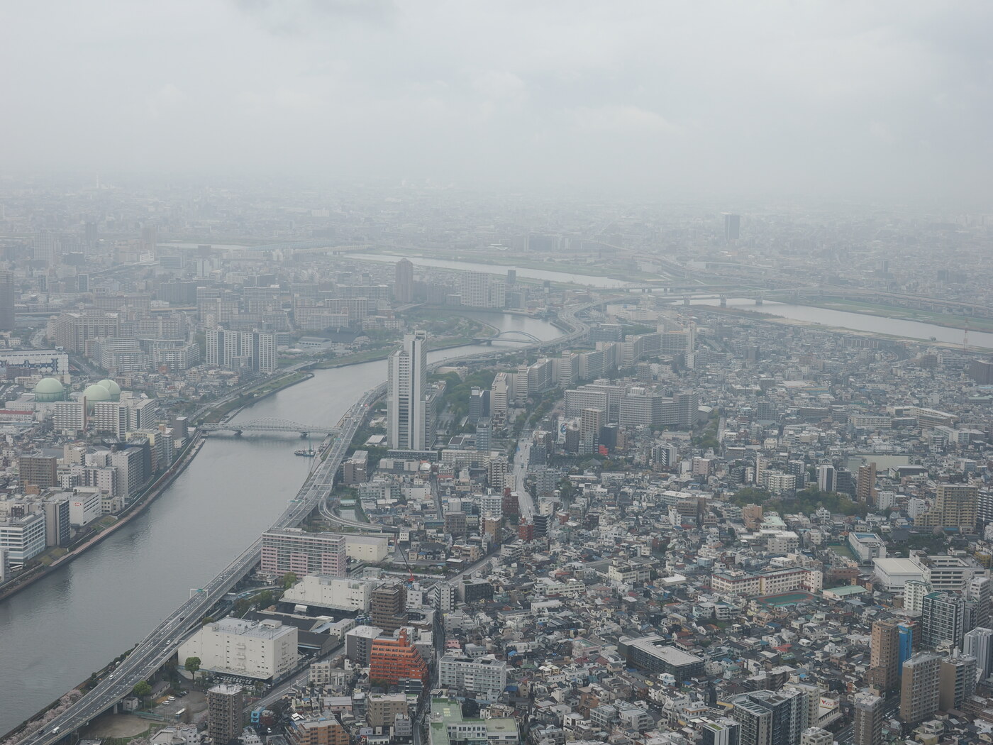 Panorama wschodnia z Tokyo Skytree — rzeka Arakawa w tle, szerokie rozlewisko, mgła nad dalszą częścią Tokio