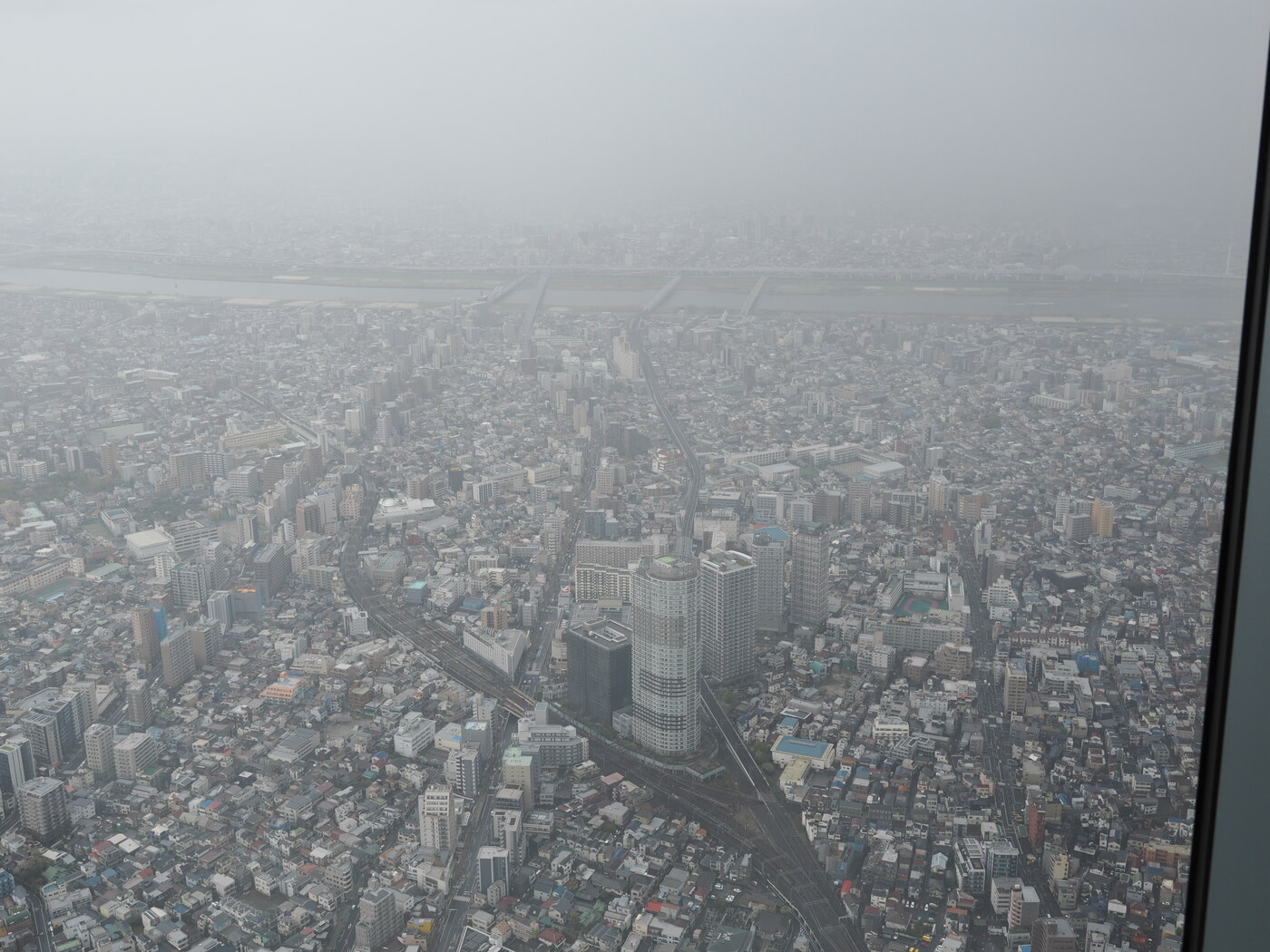 Widok na wschód z Tembo Deck — linia kolejowa JR biegnąca przez zabudowę, cylindryczny wieżowiec na środku, mgła, rzeka Arakawa w tle