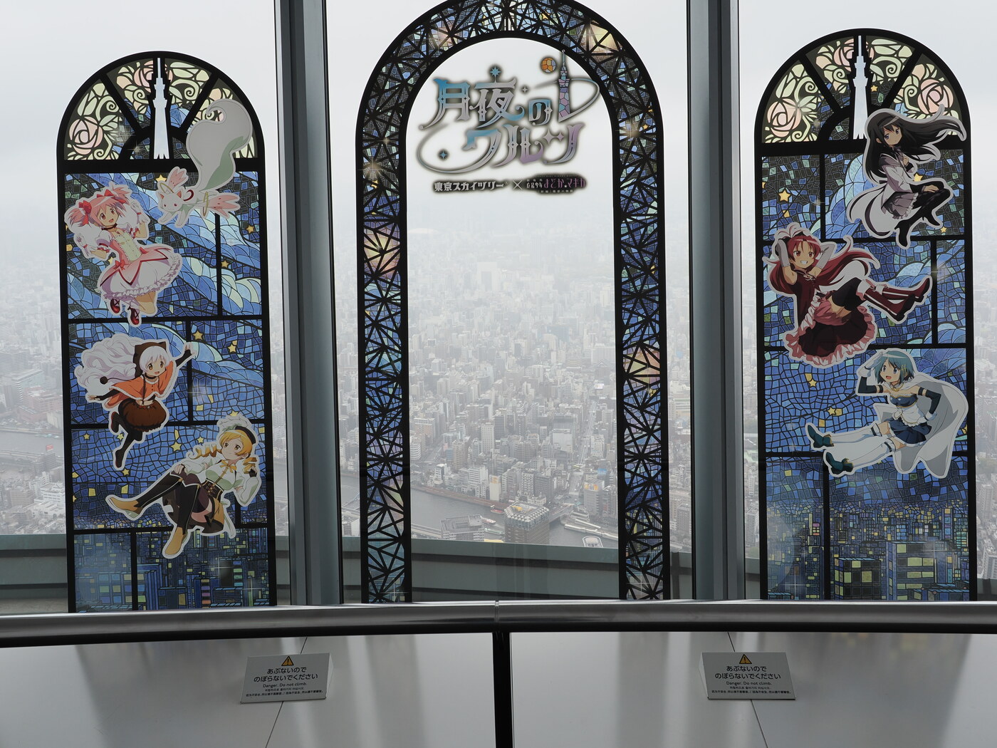 Witraż Madoka Magica × Tokyo Skytree — trzy okna witrażowe z postaciami Madoki, Nagisy, Mami i Homury unoszącymi się nad panoramą Tokio, logo 月夜のワルツ pośrodku