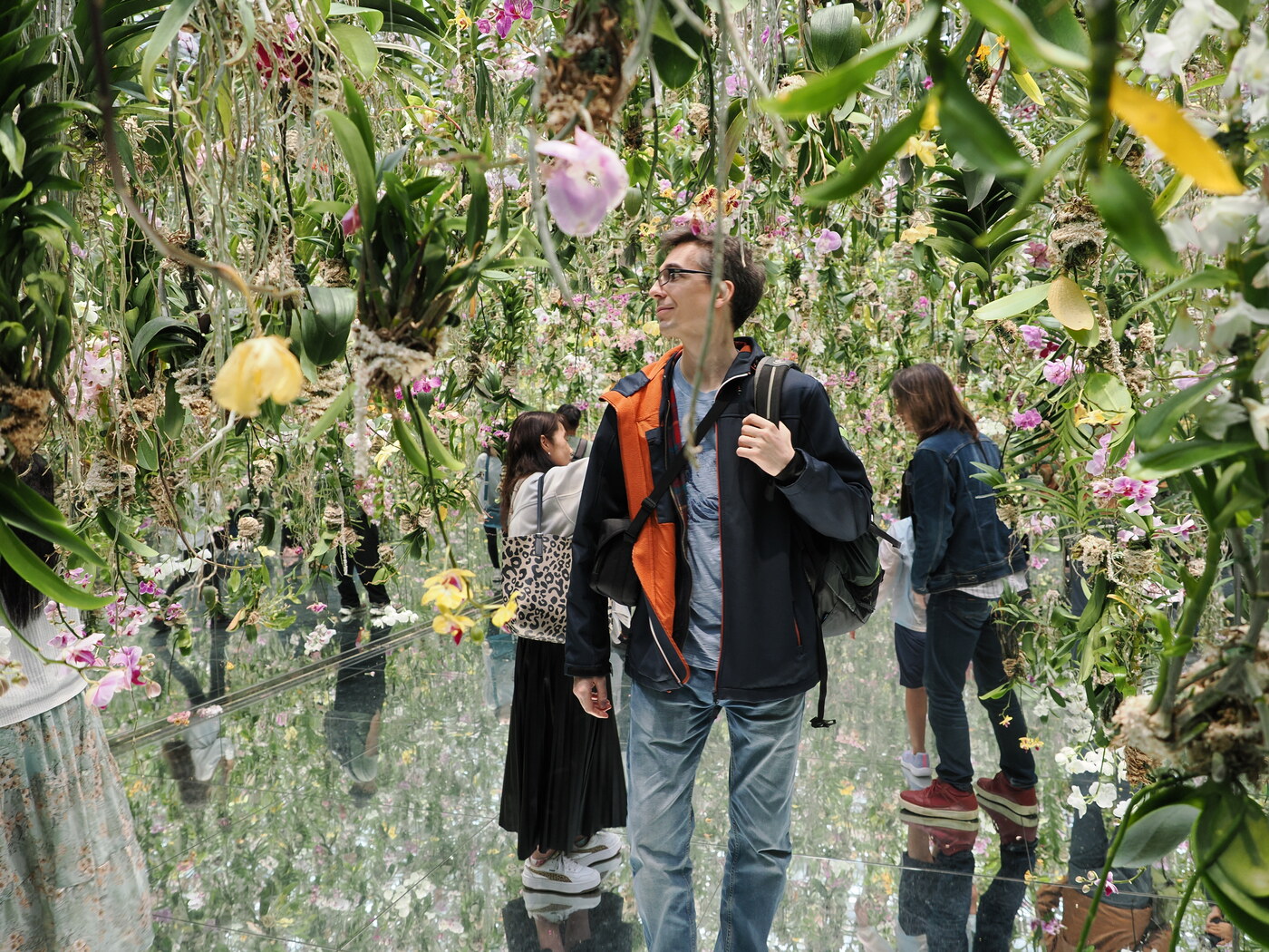 Kamil w ogrodzie orchidei teamLab Planets — mężczyzna w pomarańczowej kurtce stoi pośród zwisających orchidei i lustrzanej podłogi, różowe i żółte orchidee dookoła