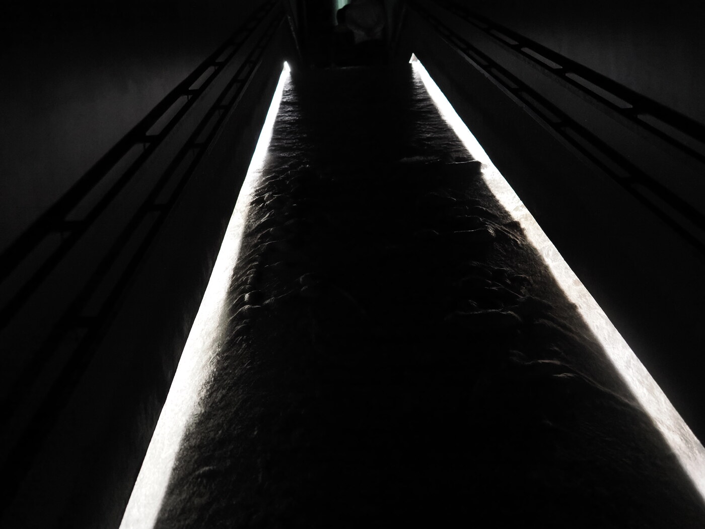 teamLab Planets — widok z dołu na wąski pionowy kanał z dwoma paskami białego światła biegnącymi ku górze, kamienne lub betonowe ściany w ciemności