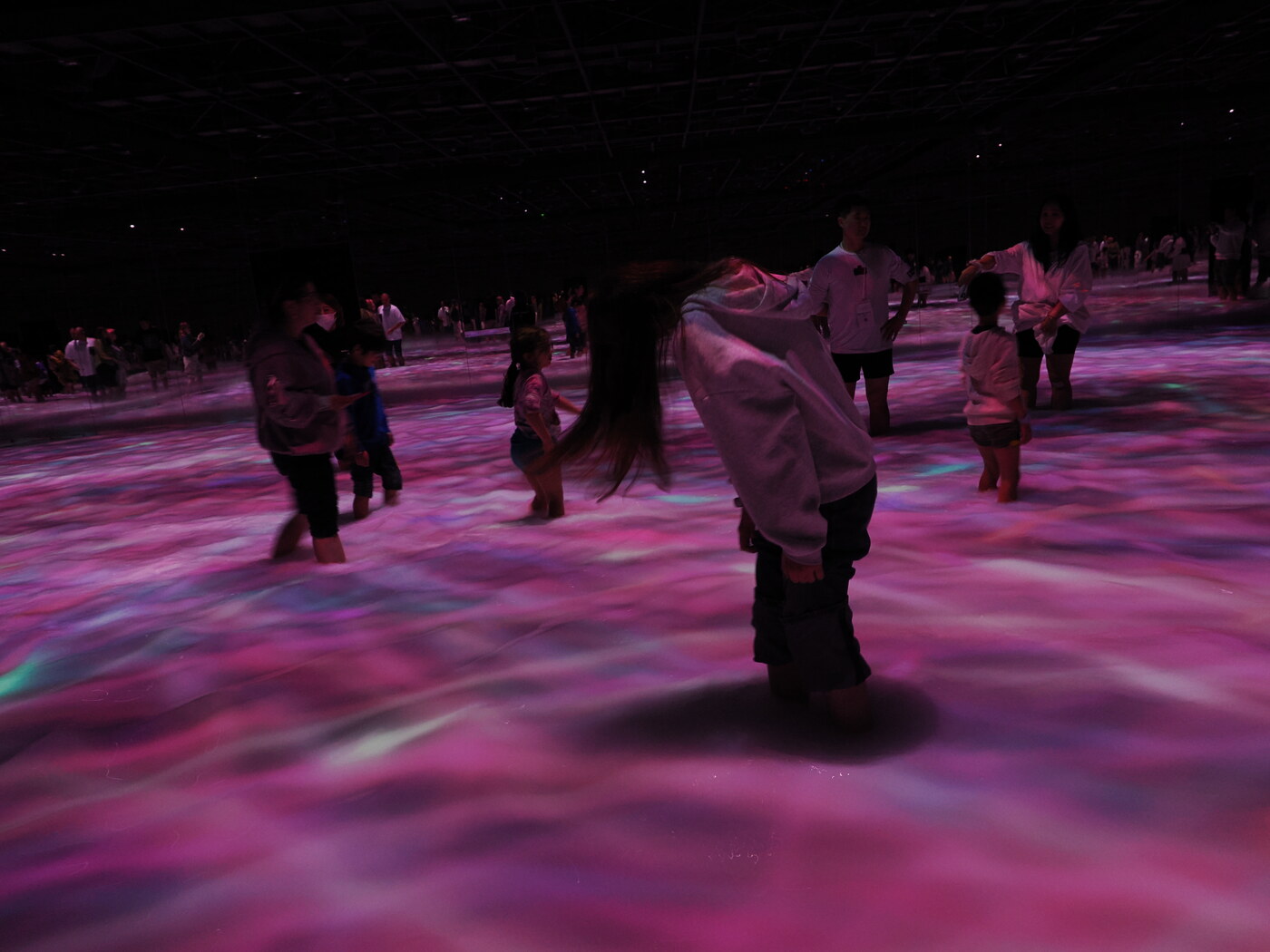 teamLab Planets — sala z projekcją na podłodze, dzieci i dorośli brodzą w różowej, falującej wodzie z projekcją zorzy polarnej, ciemny sufit