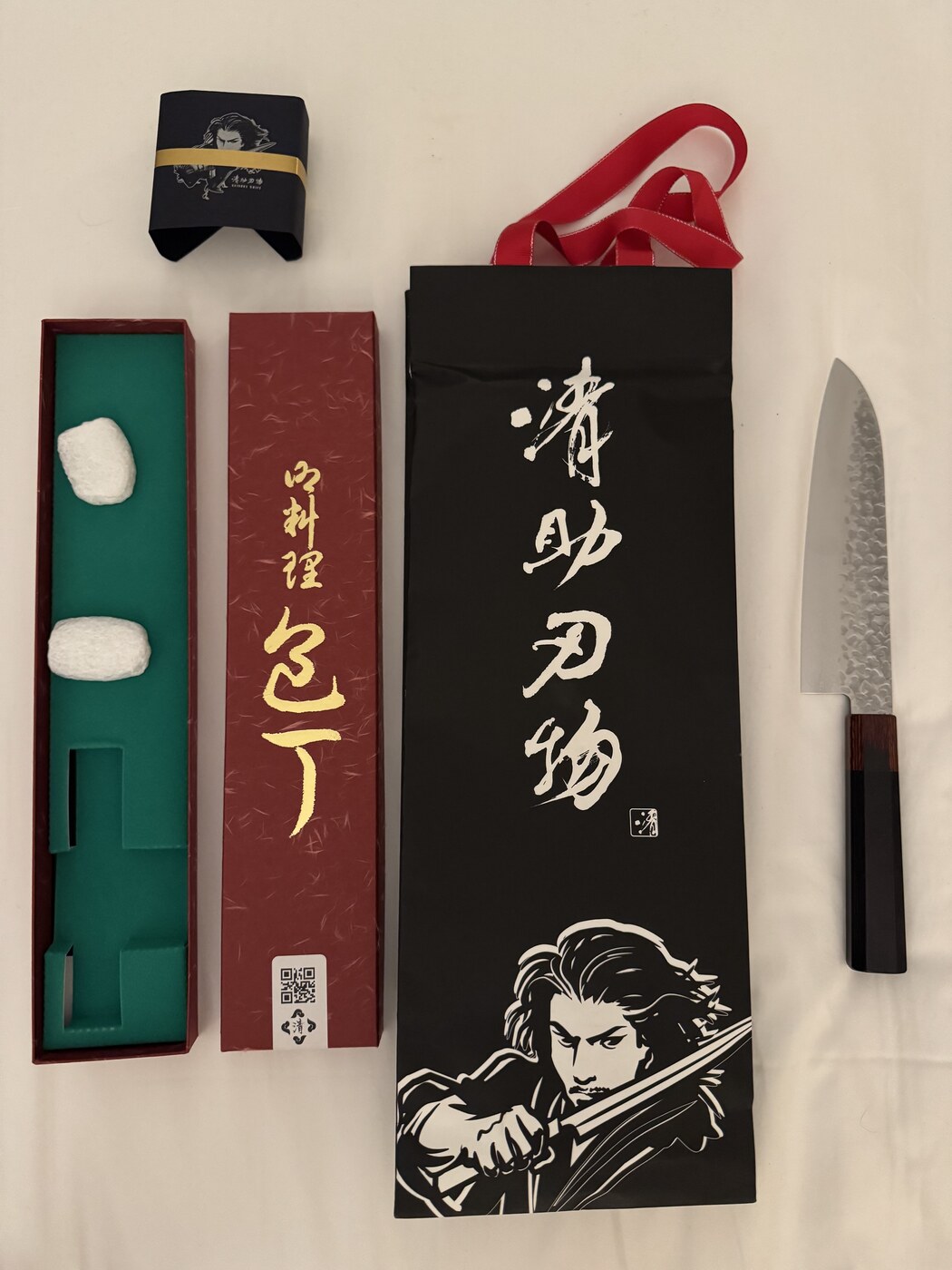 Japoński nóż santoku Seisuke z wykończeniem tsuchime w pudełku — czarna torba prezentowa z grafiką samuraja, bordowe pudełko z zielonym wnętrzem