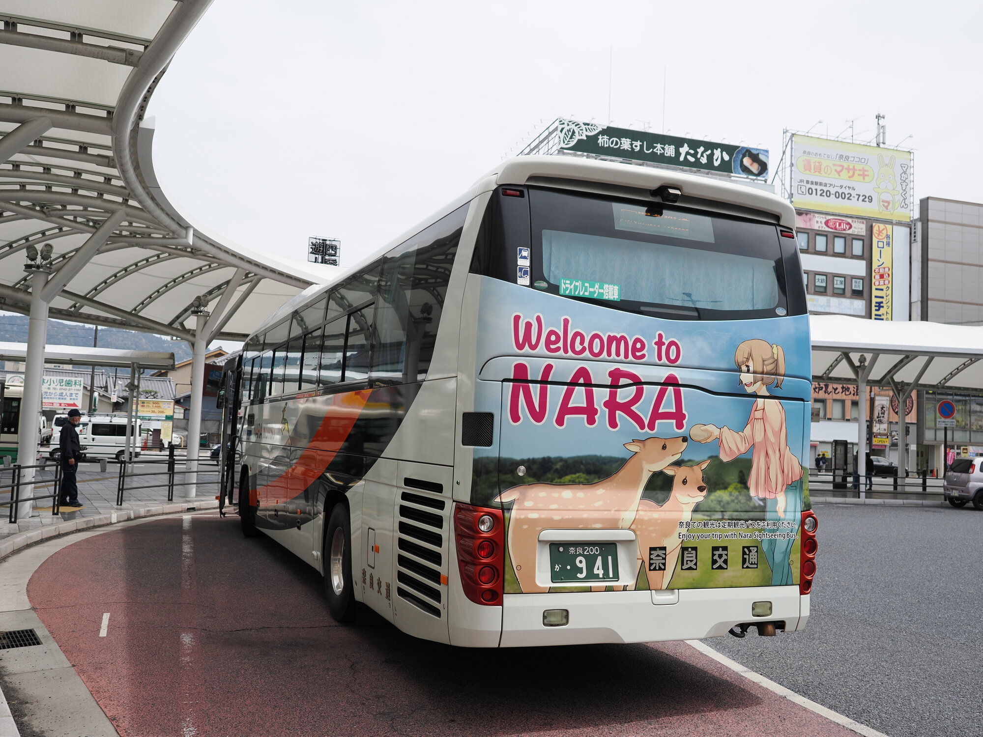 Autobus turystyczny Nara Kōtsū z napisem Welcome to NARA i grafiką anime — dziewczyna karmiąca jelenie sika, stacja JR Nara w tle