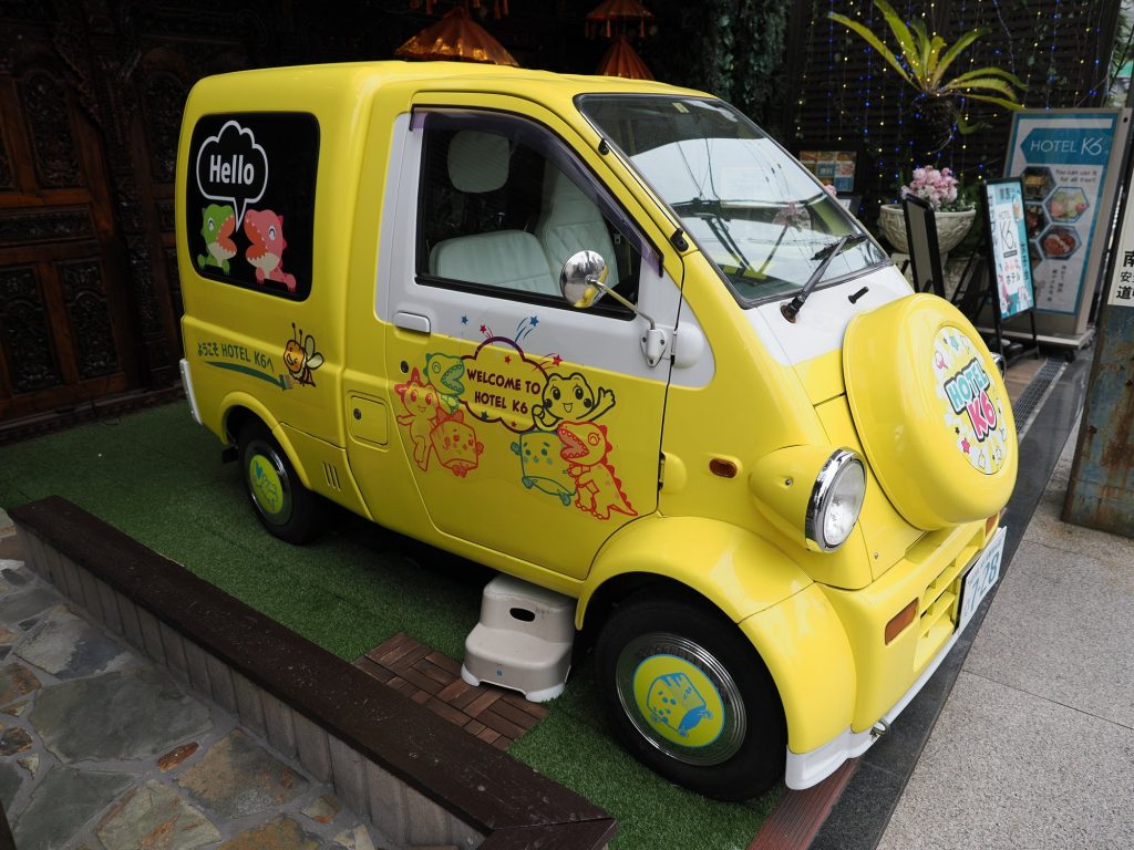 Żółty kei-van Hotel K6 z nadrukowanymi dinozaurami przed wejściem