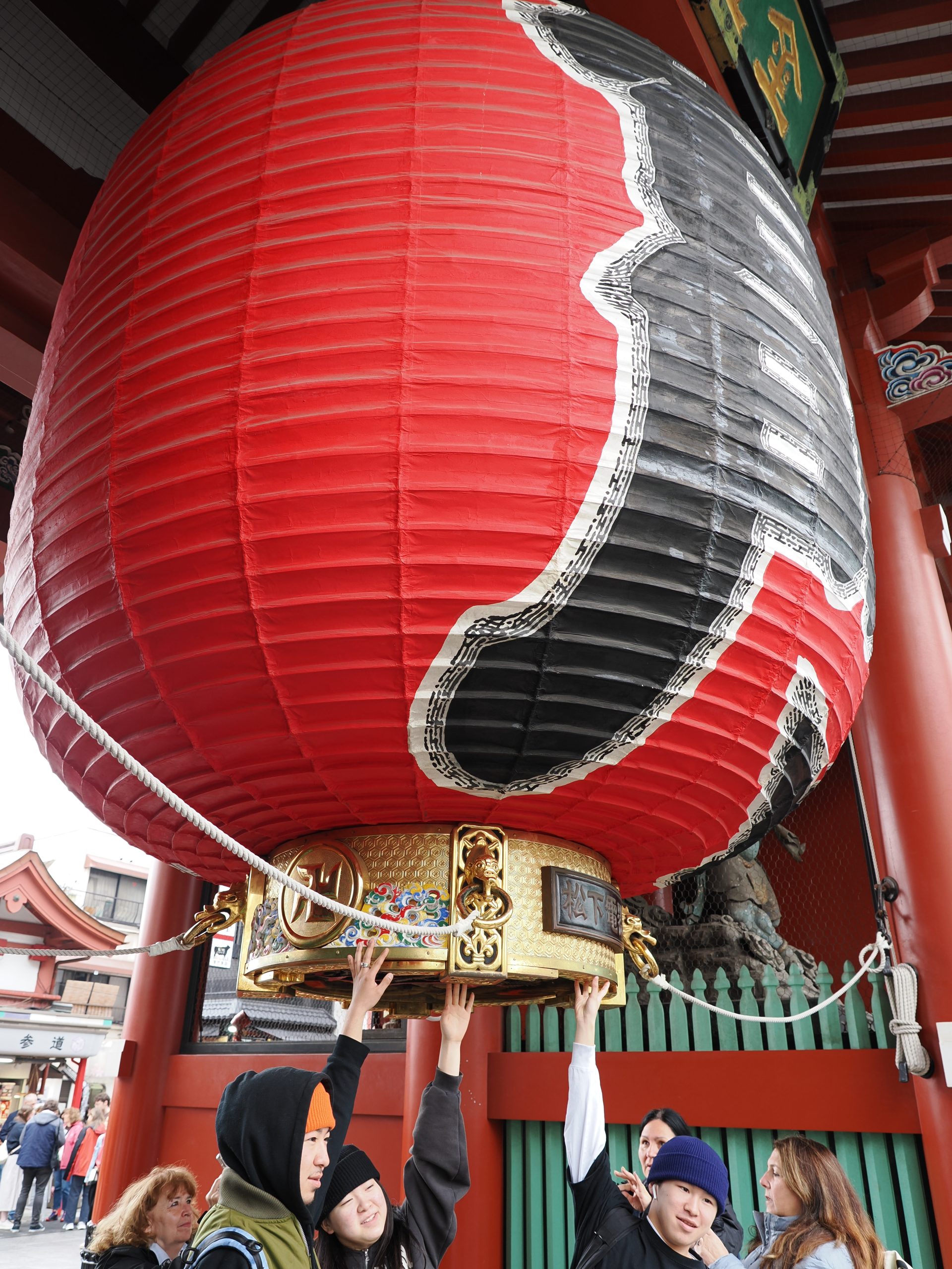 Zbliżenie na gigantyczny czerwony lampion Kaminarimon, dwoje młodych ludzi dotykających złotej podstawy