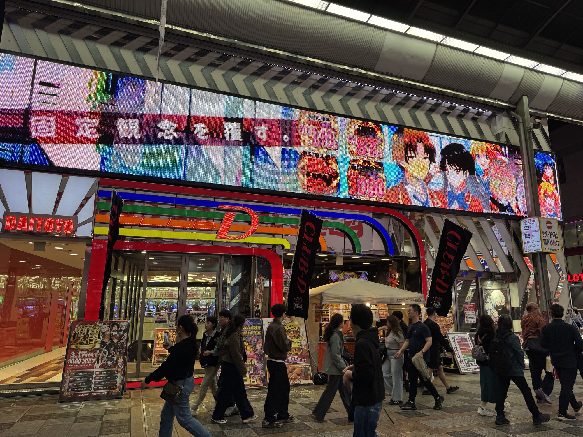 Salon pachinko w Nippombashi Osaka — jasne neony, przechodnie przy wejściu, charakterystyczna ulica rozrywki Den Den Town