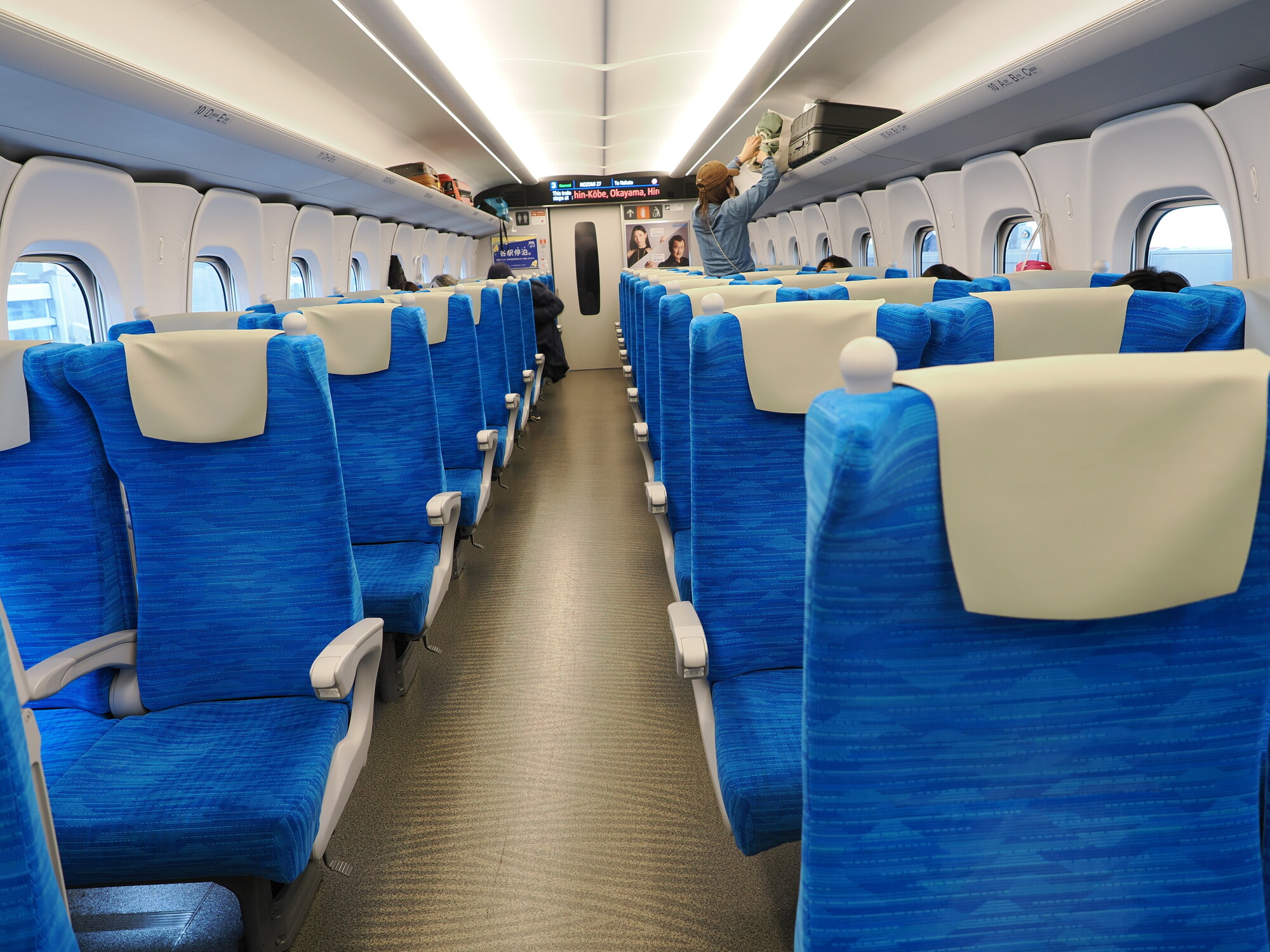 Wnętrze Shinkansena N700S — niebieskie fotele, tablice informacyjne, pasażerka wkłada bagaż na półkę, wyświetlacz Shin-Kobe Okayama