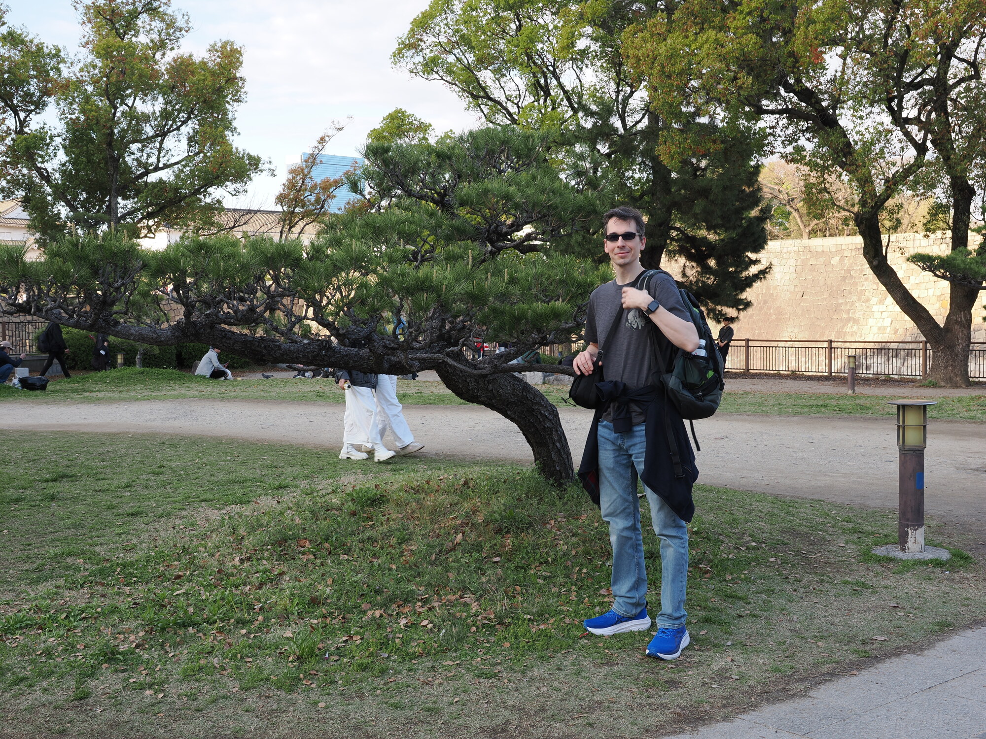 Kamil przed murami zamku Osaka — mur kamienny, sosny, latarnia ogrodowa, turyści w tle