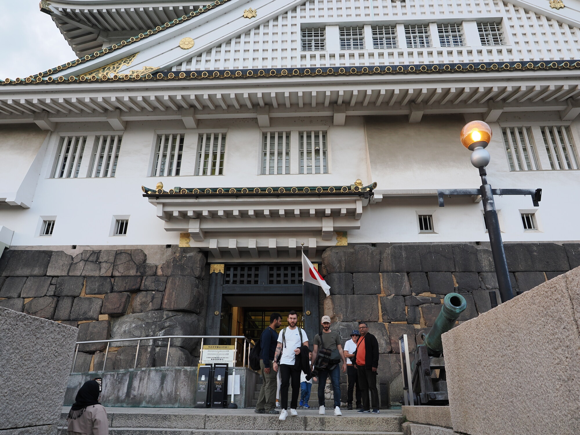 Wejście do muzeum zamku Osaka od dołu — biała fasada z czarnymi zdobieniami, kamienny fundament, działo i flaga Japonii