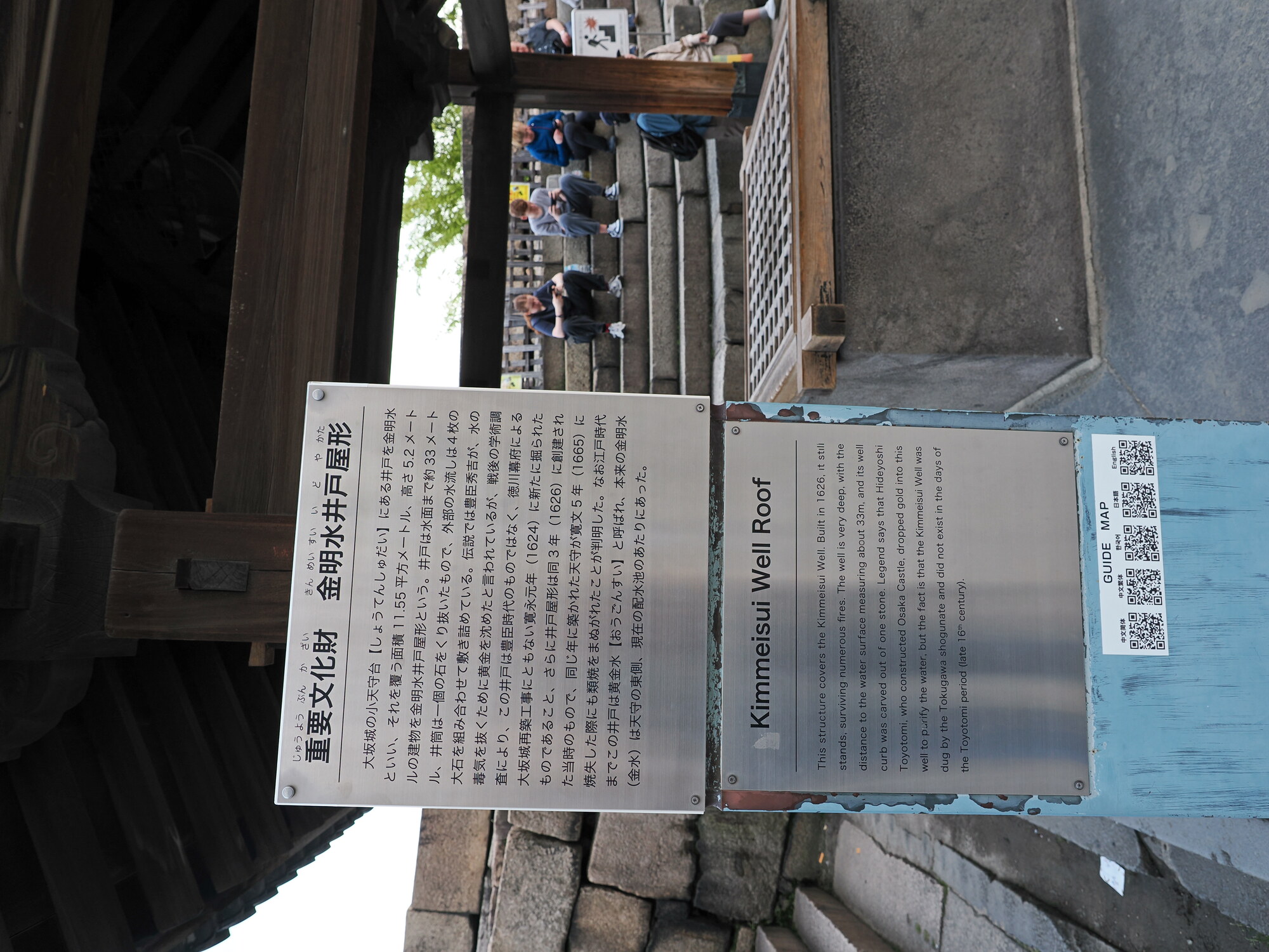 Tablica informacyjna Kimmeisui Well Roof przy zamku Osaka — opis studni z 1626 roku wykutej z jednego kamienia, turystki odpoczywają w tle