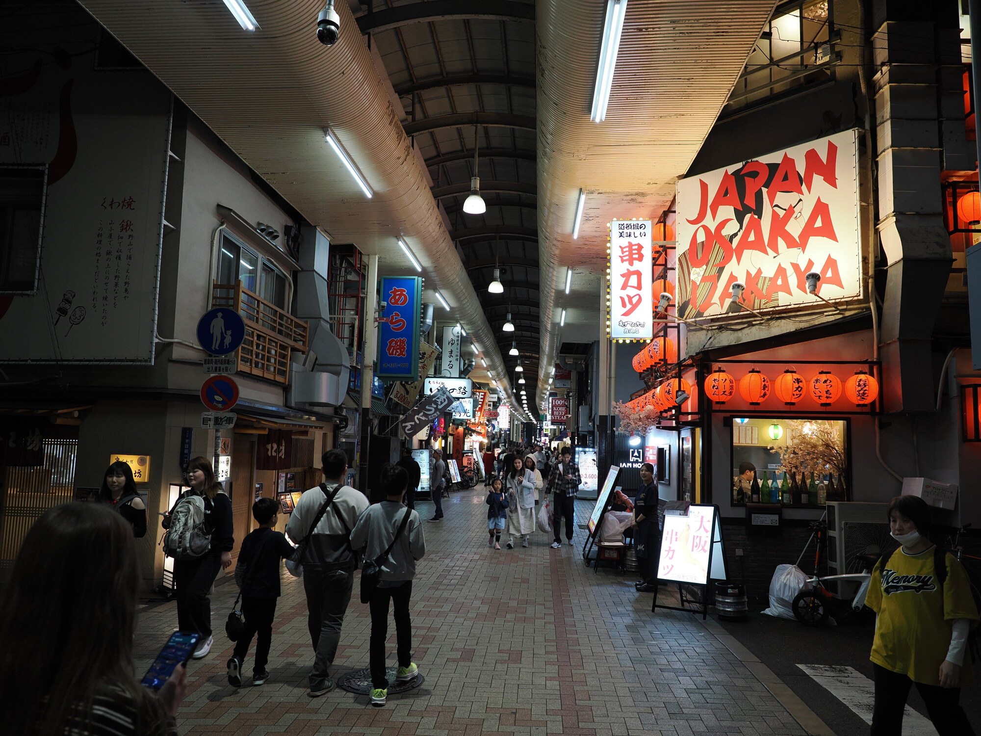 Zadaszony pasaż handlowy przy Dotonbori — sklepy kuwa-yaki i kushikatsu po bokach, szyld JAPAN OSAKA IZAKAYA po prawej