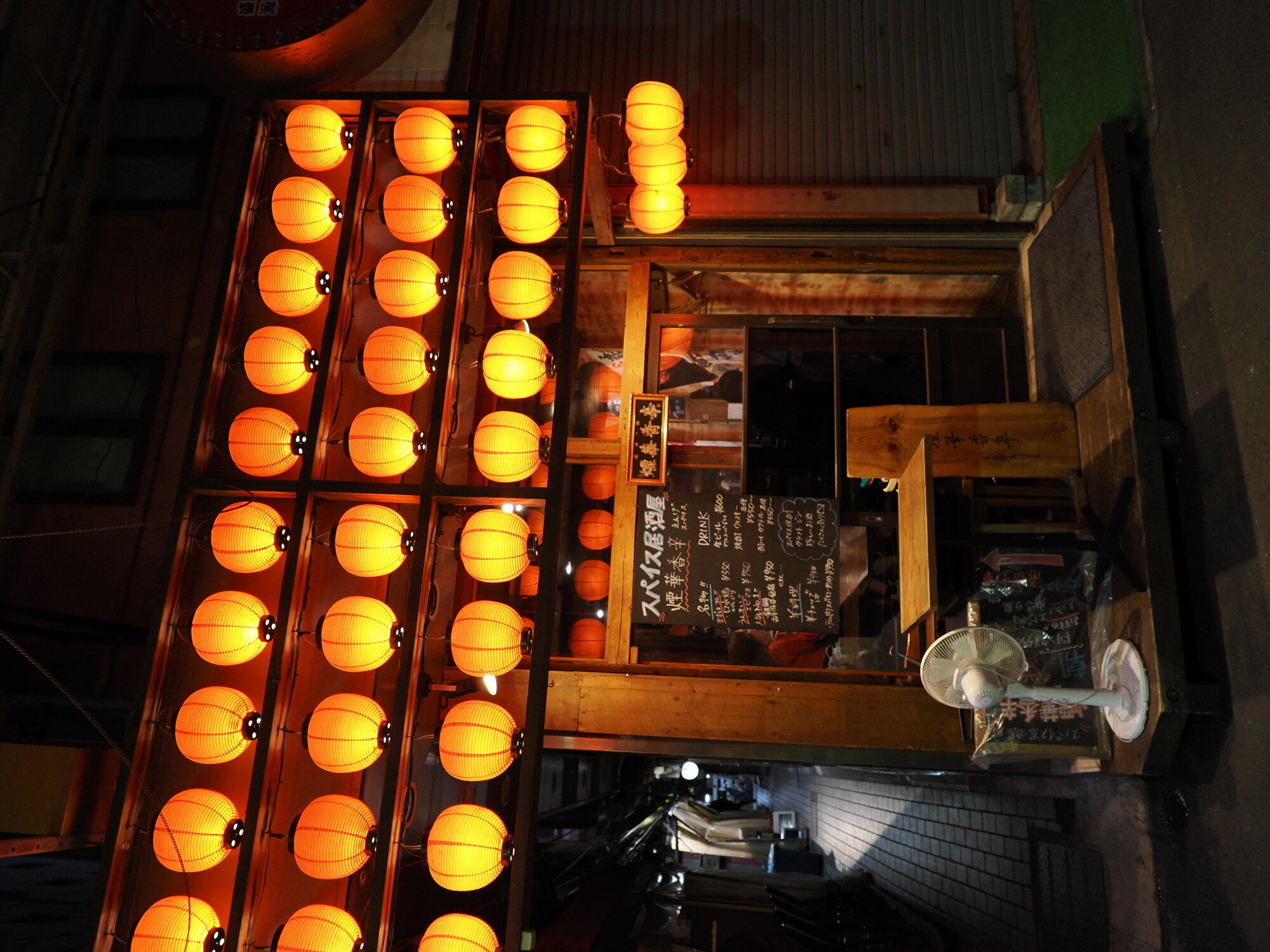 Fasada restauracji スパイス居酒屋 煙華香辛 z rzędami pomarańczowych lampionów — trzy rzędy świecących kul chochin, tablica menu na drewnianej desce