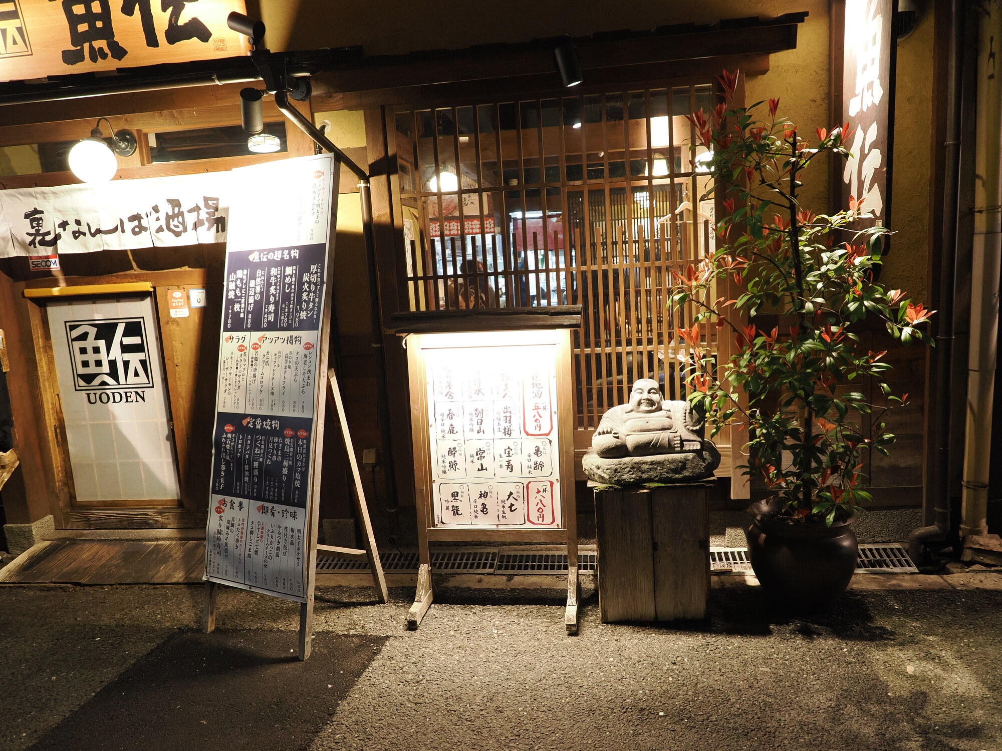 Wejście do izakayi Uoden w Namba — kamienny Hotei z wielkim brzuchem przy drzwiach, tablice menu na drewnianej desce, lampiony przy wejściu