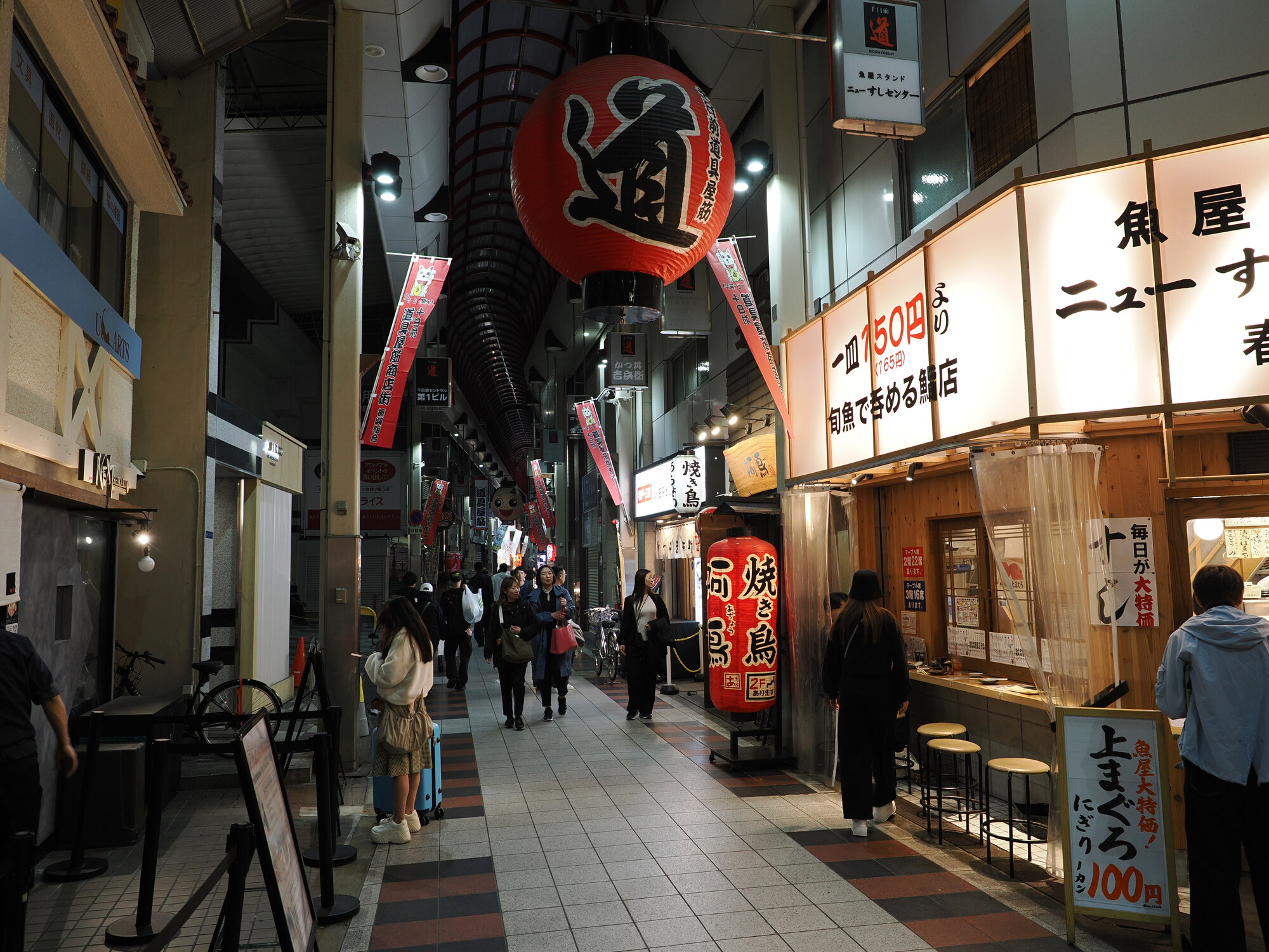 Zadaszony pasaż gastronomiczny Namba — wielki czerwony lampion 道 i bar sushi 魚屋ニュースセンター z nigiri po 100 jenów, tłum spacerujących