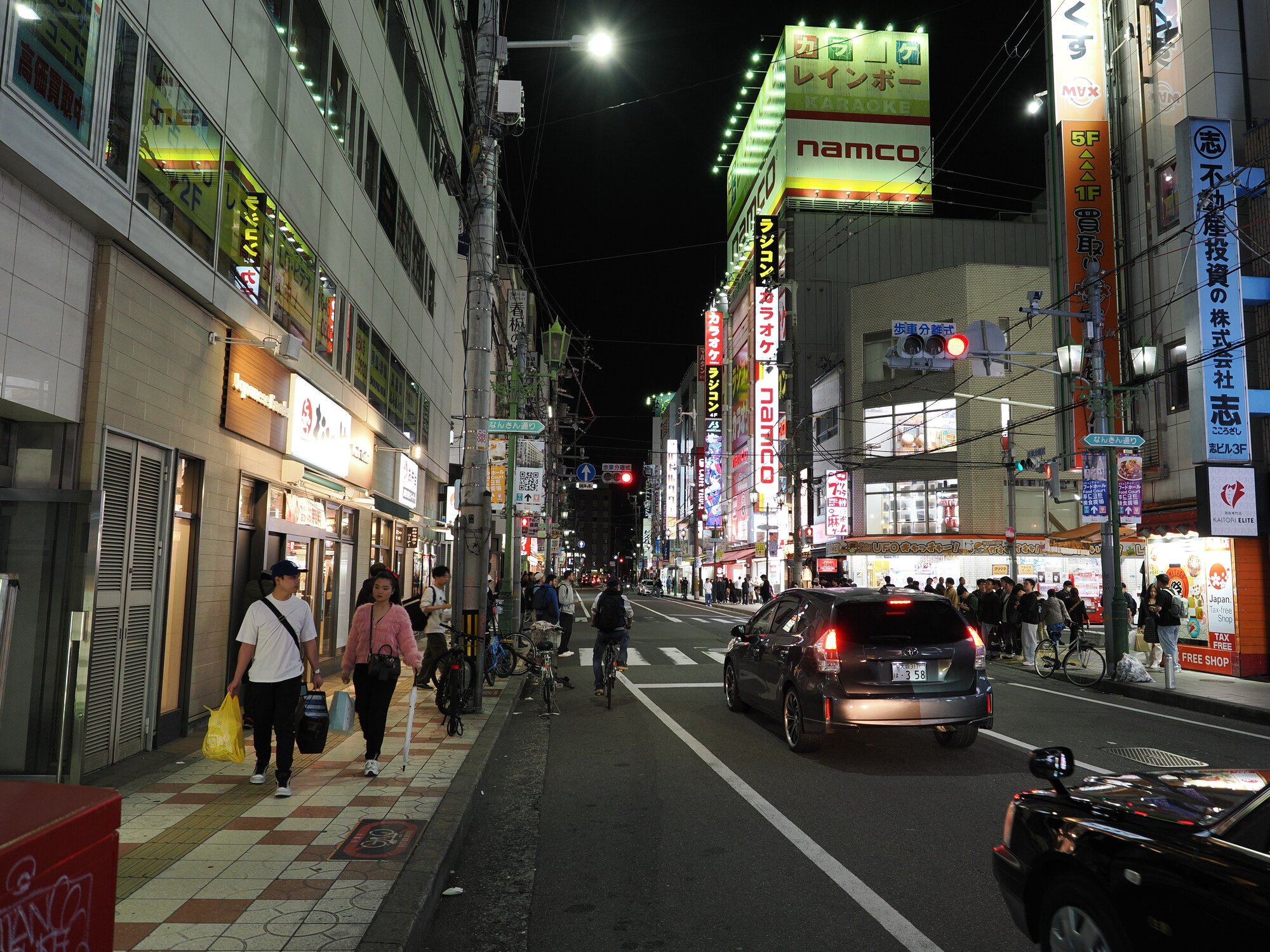 Nocna ulica Nambo — neony Namco i karaoke Rainbow, auta na jezdni, przechodnie i rowery, szyldy po obu stronach