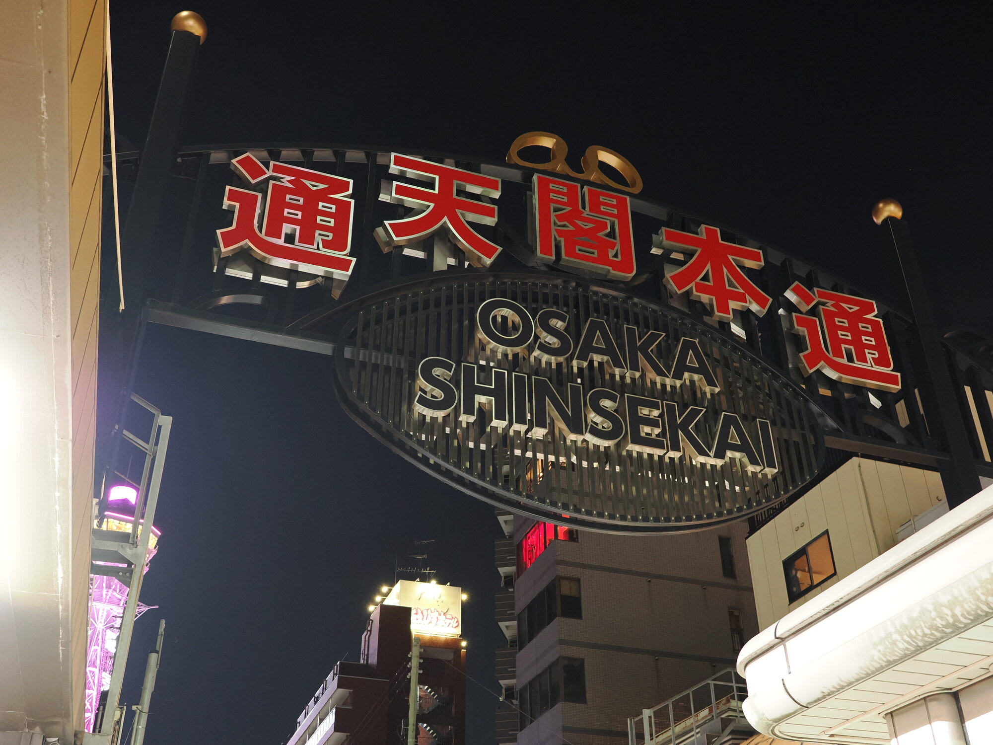 Brama Shinsekai nocą — duży szyld 通天閣本通 OSAKA SHINSEKAI z czerwonymi kanji, czarne niebo, po lewej różowe neon wieży Tsutenkaku